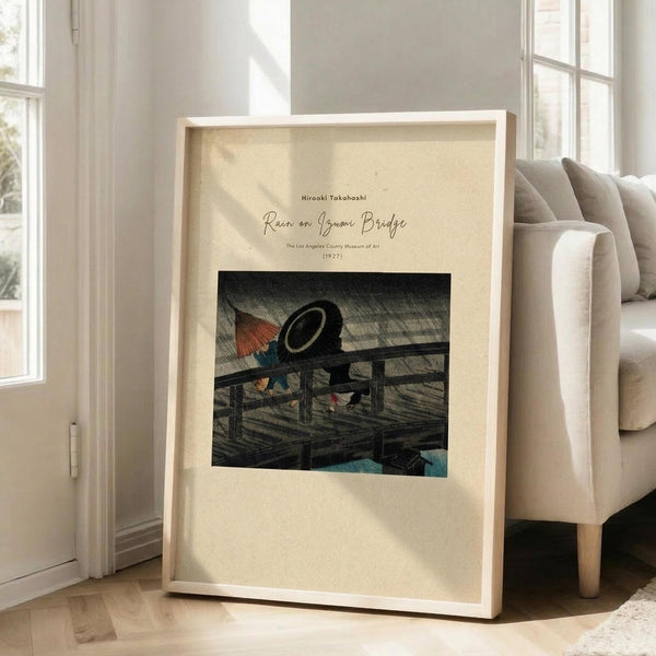 May include: Framed art print leaning against white wall next to beige upholstered chair in bright room with large windows. Print features Japanese woodblock style artwork showing figures with umbrellas on a wooden bridge in rain, with diagonal rain lines across dark sky. Cream colored mat surrounds the artwork in natural wood frame. Sunlight streams across hardwood floors creating geometric shadows.