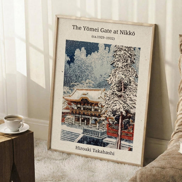 May include: Framed vintage Japanese woodblock print showing a traditional temple gate covered in snow with ornate red and gold architecture, surrounded by snow-laden trees against a dark blue sky with falling snowflakes. The artwork is displayed in a light wooden frame leaning against white wainscoting beside a coffee cup on a wooden block and cream-colored blanket on a textured white rug.