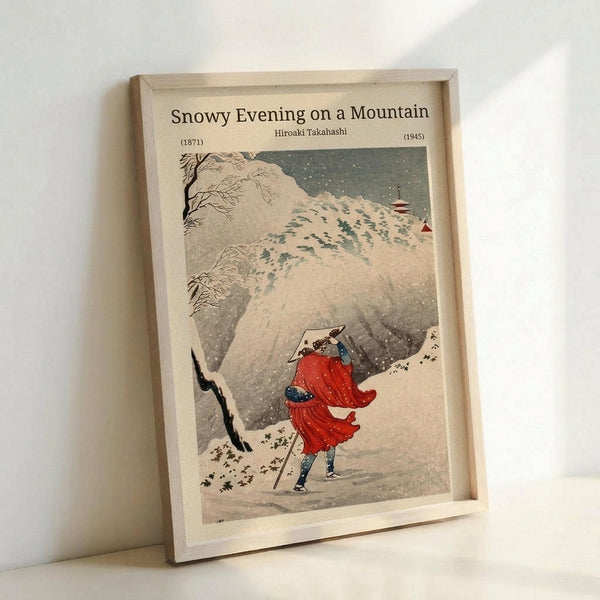 May include: Framed Japanese woodblock print reproduction showing a figure in flowing red robes walking through heavy snowfall on a mountain path. The artwork features muted earth tones with white snow against dark rocky cliffs and bare trees. The print is displayed in a light wooden frame leaning against a white wall, showing the traditional Asian art style with falling snow depicted as white dots across the composition.