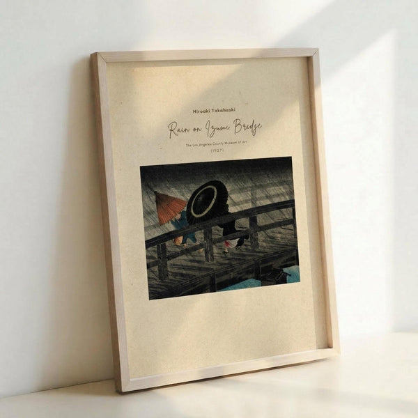May include: Framed vintage Japanese woodblock print showing people with umbrellas crossing a wooden bridge in heavy rain, depicted with diagonal rain lines. The artwork features muted earth tones with touches of red and blue against a cream-colored mat. The light wooden frame leans against a white wall on what appears to be a light surface, creating soft shadows in natural lighting.
