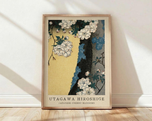 May include: Framed art print featuring Japanese cherry blossoms with white flowers against yellow and blue background in traditional woodblock style, displayed in light wood frame leaning against white textured wall on light wooden floor with soft natural lighting and shadows