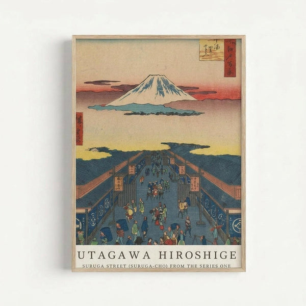 A framed Japanese woodblock print showing Mount Fuji in the background with snow-capped peak against pink and orange sky. The foreground depicts a busy street scene with many small figures in colorful traditional clothing walking through what appears to be a covered marketplace or shopping district with blue-tiled roofs. The artwork features the characteristic style of ukiyo-e prints with flat colors, detailed architectural elements, and Japanese text in the upper right corner. The print is disp