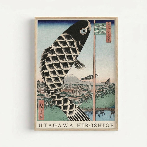 Japanese woodblock print featuring a large black carp kite with detailed scale patterns swimming upward against a pale blue sky. Below lies a scenic landscape with Mount Fuji in the distance, traditional buildings, green fields, and a wooden bridge crossing a river. Additional smaller carp kites float in the background. The artwork displays classic ukiyo-e style with muted colors and includes red Japanese text cartouches. Presented in a light wooden frame with "UTAGAWA HIROSHIGE" text at the bot
