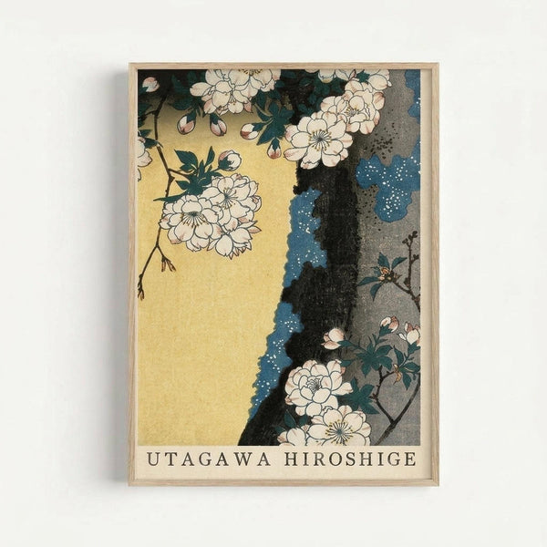 Framed Japanese woodblock print featuring delicate white cherry blossoms on dark branches against a golden yellow background. The composition includes blue and gray rocky elements with small white dots suggesting texture or moss. The artwork displays traditional ukiyo-e style with fine black line work and muted color palette. Text "UTAGAWA HIROSHIGE" appears at bottom in black letters. Natural wood frame surrounds the print, displayed against a light gray wall.