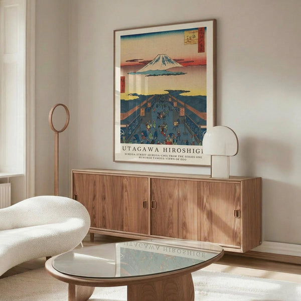 May include: Japanese woodblock print artwork displayed above wooden credenza in modern living room. The framed poster shows Mount Fuji in upper panel and busy street scene with figures in traditional dress in lower panel, featuring blue, red, and yellow colors. White curved chair and glass coffee table visible in foreground.