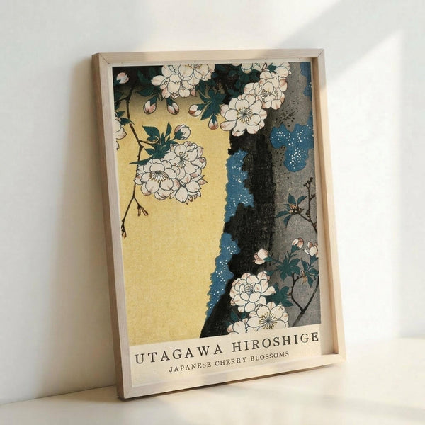 May include: Framed art print featuring Japanese cherry blossoms with white flowers on dark branches against a golden yellow background and blue water with white dots. The artwork shows traditional Japanese woodblock print style with delicate line work. Light wooden frame displayed leaning against white wall, casting soft shadows.
