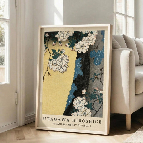 May include: A framed Japanese woodblock print featuring white cherry blossoms against a yellow and blue background with a dark tree trunk. The artwork shows delicate floral details in traditional ukiyo-e style. The print is displayed in a light wooden frame, leaning against a beige upholstered chair in a bright room with white walls and windows. Soft natural lighting creates shadows on the hardwood floor.