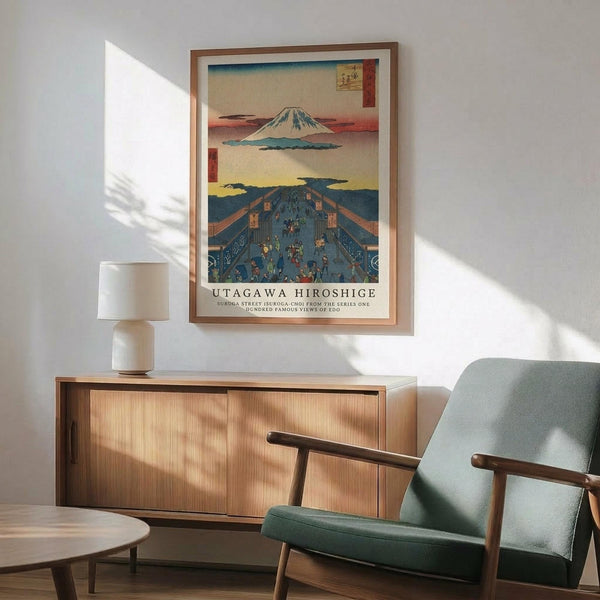 May include: Japanese woodblock print poster featuring Mount Fuji and traditional street scene with crowds of people, displayed in thin brown frame on white wall above wooden credenza. Mid-century modern room setting with sage green upholstered chair, white ceramic table lamp, and natural sunlight creating geometric shadows across the space.