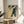 May include: A framed art print featuring white cherry blossoms against a yellow sky and dark blue tree trunk, displayed on a light wooden round side table next to a beige upholstered chair. Natural sunlight streams across white walls, with a green plant visible in the corner. The artwork shows traditional Japanese woodblock print styling with delicate floral details.