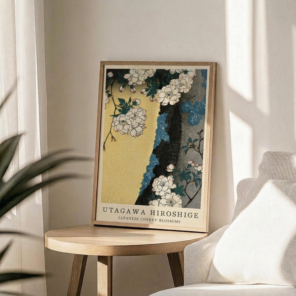 May include: A framed art print featuring white cherry blossoms against a yellow sky and dark blue tree trunk, displayed on a light wooden round side table next to a beige upholstered chair. Natural sunlight streams across white walls, with a green plant visible in the corner. The artwork shows traditional Japanese woodblock print styling with delicate floral details.