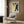 May include: Framed poster print featuring Japanese cherry blossoms in traditional ukiyo-e style with white flowers, dark branches, yellow and blue background, displayed on wood-paneled wall above beige upholstered chair with geometric throw pillow, brass floor lamp and side table with open book nearby