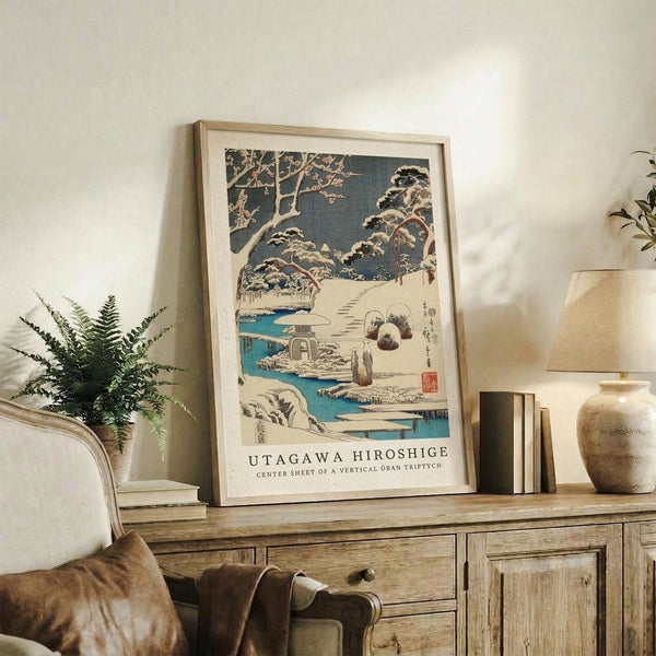 May include: Framed Japanese woodblock print featuring winter landscape with snow-covered trees, blue water, stone lantern, and figures in traditional dress, displayed on rustic wooden sideboard alongside fern plant, ceramic vase, table lamp, and books in warm-toned living room setting