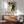May include: Framed Japanese woodblock print featuring cherry blossoms with white flowers against yellow sky and dark tree trunk, displayed above mid-century modern wooden sideboard with white ceramic lamp, next to sage green upholstered armchair with wooden frame in sunlit room with white walls