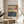 May include: Framed Japanese woodblock print reproduction featuring Mount Fuji in background and busy blue street scene with traditional figures below, displayed on light wood round side table next to beige upholstered chair. Natural lighting creates soft shadows on white wall. Green plant leaves visible in corner of minimalist room setting.
