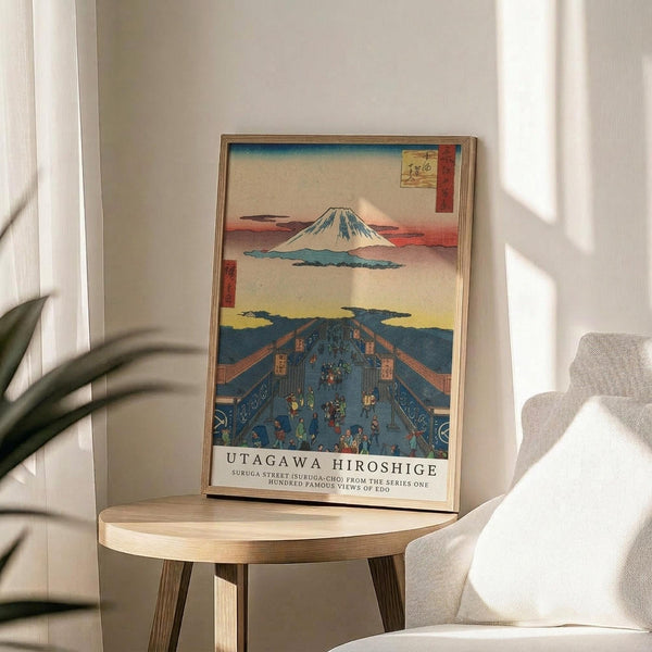 May include: Framed Japanese woodblock print reproduction featuring Mount Fuji in background and busy blue street scene with traditional figures below, displayed on light wood round side table next to beige upholstered chair. Natural lighting creates soft shadows on white wall. Green plant leaves visible in corner of minimalist room setting.