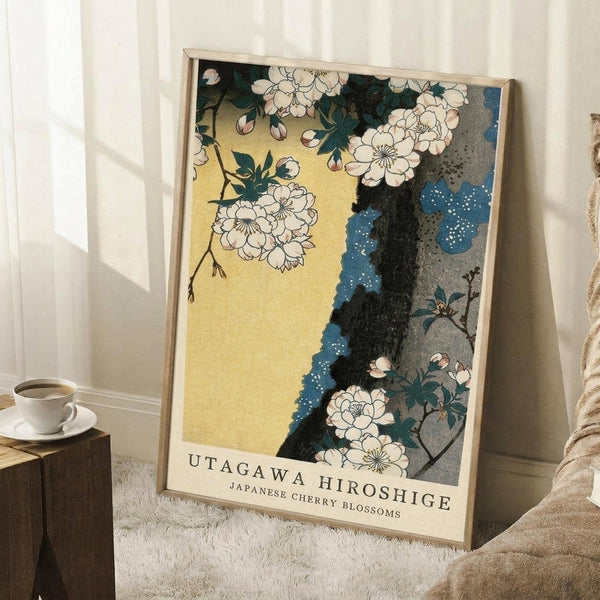May include: A framed poster featuring Japanese cherry blossoms with white flowers on dark branches against a yellow and blue background with decorative dots, displayed in a light wood frame leaning against white walls in a cozy room setting with a coffee cup on a wooden side table and beige textured throw blanket nearby.