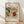 May include: Framed abstract art print leaning against white wall on light wood floor. Colorful expressionist painting features swirling brushstrokes in vibrant yellows, reds, blues, and greens with organic flowing forms. Light wood frame contains cream matting with black text above and below the artwork. Soft natural lighting creates diagonal shadows across the scene.