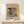 May include: Framed abstract art print featuring vibrant swirling colors in blues, yellows, reds, and greens with dynamic brushstrokes and geometric elements. Print displays beige background with cursive script and museum attribution text. Wooden frame sits on light wood surface beside small white wrapped gift with evergreen sprig against cream wall.
