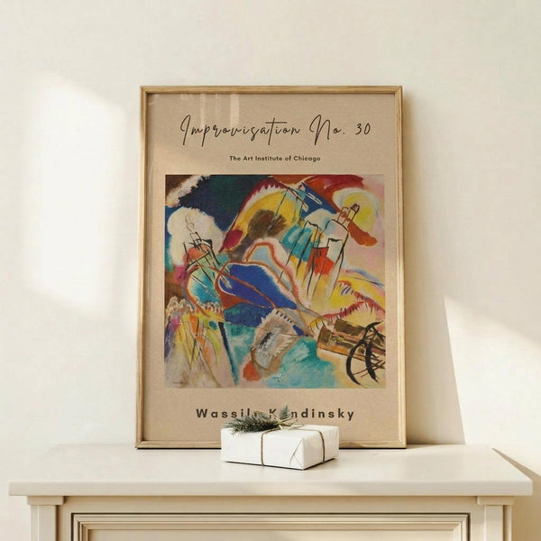 May include: Framed abstract art print featuring vibrant swirling colors in blues, yellows, reds, and greens with dynamic brushstrokes and geometric elements. Print displays beige background with cursive script and museum attribution text. Wooden frame sits on light wood surface beside small white wrapped gift with evergreen sprig against cream wall.