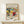 May include: Abstract expressionist artwork poster in wooden frame displayed on light wood console table against cream wall. Vibrant painting features flowing organic forms in blues, yellows, reds, and greens with dynamic brushstrokes and curved lines. Small white wrapped gift with natural twine and evergreen sprig sits beside the framed print. Warm, minimalist home decor setting with soft natural lighting.