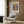 May include: Framed Claude Monet poster print hanging on wood-paneled wall above beige upholstered armchair. Artwork shows impressionist sunrise scene with orange sun reflecting on blue water, boats silhouetted against colorful sky. Modern living room setting with brass floor lamp, geometric throw pillow, and dark side table with open book.
