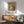 May include: Framed art print of Monet's sunrise seascape hanging above wooden sideboard in modern living room. Print shows golden sunset over ocean with solitary figure on beach, displayed in thin wooden frame. Mint green mid-century armchair and white ceramic table lamp visible, with warm natural lighting creating shadows on white wall.