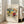 May include: Framed abstract art print leaning against white wall next to beige sofa in bright living room. Colorful expressionist painting features bold brushstrokes in yellows, blues, greens, and reds with fluid organic forms. Light wood frame with white mat displays the vibrant artwork on hardwood floors near large windows.