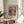 May include: Framed poster print of impressionist sunrise seascape with orange sun reflecting on blue water, boats silhouetted against colorful sky. Features Claude Monet signature and quote below artwork. Gold-toned frame displayed on round wooden side table next to beige upholstered chair with white throw pillow. Natural sunlight creates shadows on neutral wall background with plant leaves visible in corner.