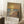 May include: Framed art print leaning against white wall showing impressionist-style seascape with golden sunset, blue ocean waves, sandy beach, and solitary figure walking. Warm yellow and orange sky reflects on water. Text reads "MONET" at bottom. Print has light wood frame, positioned on white textured rug near coffee cup on wooden surface and beige fabric.
