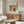 May include: Abstract colorful art print displayed above wooden sideboard in modern living room setting. Vibrant painting features bold brushstrokes in yellow, red, blue, and green with organic flowing forms. Print shown in thin wooden frame on neutral wall. Room includes curved white textured chair, glass oval coffee table, brass floor lamp, and white table lamp on sideboard.
