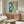 May include: Abstract watercolor artwork in turquoise, teal and cream tones with organic flowing shapes and splatter effects, displayed above a mid-century modern walnut credenza in a minimalist living room with white curved chair, glass coffee table, and brass floor lamp