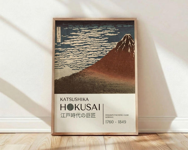 May include: Framed vintage Japanese woodblock print showing Mount Fuji in reddish-brown tones against blue sky with white clouds, displayed in light wood frame leaning against white wall on wooden floor. Sunlight creates diagonal shadows across the scene. Print features Japanese text and dates 1760-1849 below the artwork.