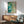 May include: Abstract watercolor artwork in turquoise, teal, and cream tones with organic flowing shapes and scattered dots, displayed in thin wooden frame above mid-century modern credenza. Sage green upholstered armchair with wooden arms sits nearby alongside small round side table and white ceramic table lamp. Warm natural sunlight streams across white wall creating geometric shadows.