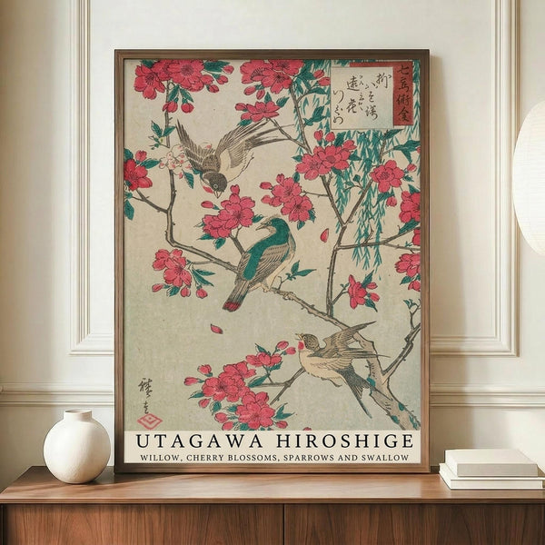 May include: A framed reproduction of a traditional Japanese woodblock print showing cherry blossoms and willow branches with sparrows and a swallow. The artwork features pink flowers, green foliage, and birds in classic ukiyo-e style with Japanese text. The print is displayed in a thin brown frame on a wooden credenza alongside a white ceramic vase and stacked books in a bright, modern room with white walls and molding.