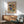 May include: A framed abstract expressionist poster with vibrant splashes of blue, yellow, red, and green against a cream background, displayed above a mid-century modern wood sideboard. The artwork features flowing organic shapes and bold brushstrokes. Scene includes a sage green upholstered armchair with wooden frame, small round side table, and white ceramic table lamp with natural sunlight casting geometric shadows across white walls.