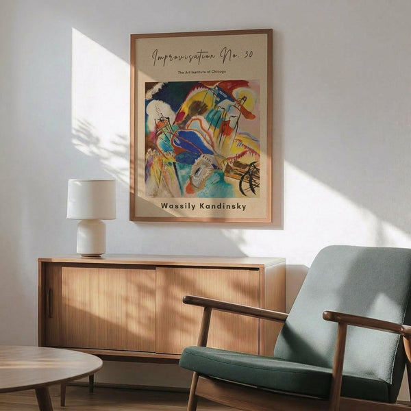 May include: A framed abstract expressionist poster with vibrant splashes of blue, yellow, red, and green against a cream background, displayed above a mid-century modern wood sideboard. The artwork features flowing organic shapes and bold brushstrokes. Scene includes a sage green upholstered armchair with wooden frame, small round side table, and white ceramic table lamp with natural sunlight casting geometric shadows across white walls.