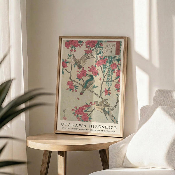 May include: Framed Japanese woodblock print featuring pink cherry blossoms and colorful birds perched on willow branches, displayed on a round wooden side table next to a beige upholstered chair. Sunlight streams across white walls creating geometric shadows, with a green plant visible in the corner. The artwork shows traditional ukiyo-e style with birds in blue, brown and muted tones among delicate spring flowers.