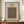 May include: Framed poster displaying an ornate carpet pattern with intricate floral and geometric designs in deep blues, oranges, and golden tones. The print features museum-style text identifying it as a William Morris design from 1834-1896. Shown in thin brown frame on wooden sideboard next to small cream ceramic vase and stacked books against cream paneled wall.
