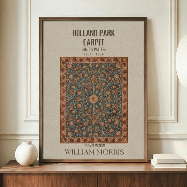 May include: Framed poster displaying an ornate carpet pattern with intricate floral and geometric designs in deep blues, oranges, and golden tones. The print features museum-style text identifying it as a William Morris design from 1834-1896. Shown in thin brown frame on wooden sideboard next to small cream ceramic vase and stacked books against cream paneled wall.
