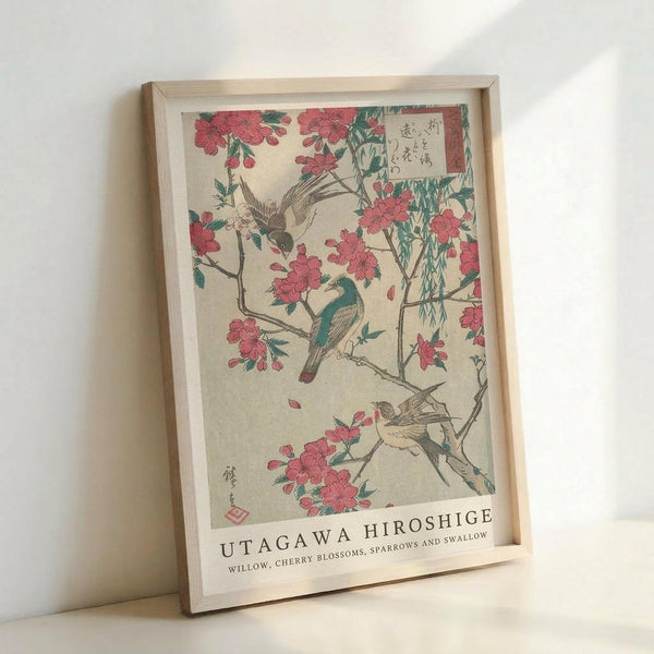 May include: Framed Japanese woodblock print featuring pink cherry blossoms and various birds including sparrows and a swallow perched on flowering branches with willow. The artwork displays traditional ukiyo-e style with muted colors on cream background, Japanese text in upper corner, and artist attribution "Utagawa Hiroshige" at bottom. Natural wood frame leans against white wall.