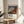 May include: Framed art print featuring Hokusai's iconic Mount Fuji with reddish-brown slopes against blue sky filled with white clouds, displayed on light wood round side table next to beige upholstered chair with white throw pillow, green plant leaves visible on left, natural sunlight casting shadows on white wall