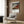 May include: Framed Japanese woodblock print poster featuring Mount Fuji in reddish-brown tones against a dark sky with white clouds, displayed on wood-paneled wall above beige upholstered armchair with geometric patterned pillow, next to brass floor lamp and dark round side table with open book