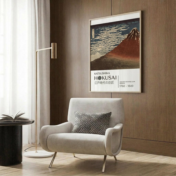 May include: Framed Japanese woodblock print poster featuring Mount Fuji in reddish-brown tones against a dark sky with white clouds, displayed on wood-paneled wall above beige upholstered armchair with geometric patterned pillow, next to brass floor lamp and dark round side table with open book