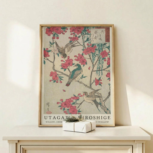 May include: Japanese woodblock print style artwork in light wood frame on white wooden dresser. Features pink cherry blossoms, small birds including green and brown swallow, with Japanese text. White gift box sits in front of frame. Print shows delicate floral branches against cream background in traditional ukiyo-e art style.