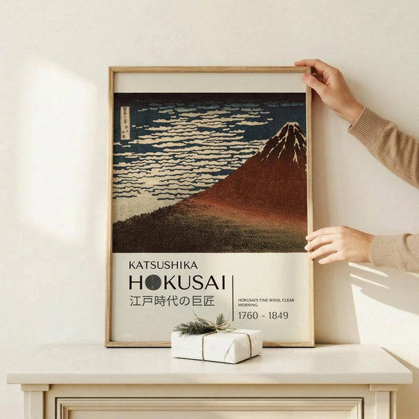 May include: Hands positioning a framed Japanese woodblock print showing Mount Fuji in reddish-brown tones against a dark sky with white clouds. The artwork features Japanese text and dates 1760-1849. A small white wrapped gift with twine sits on a light wooden surface below the light wood frame.
