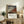 May include: Framed Japanese woodblock print featuring Mount Fuji in reddish-brown tones against blue sky with white clouds, displayed on rustic wooden sideboard alongside potted fern, ceramic vase, and stacked books in neutral-toned living room setting with beige armchair visible
