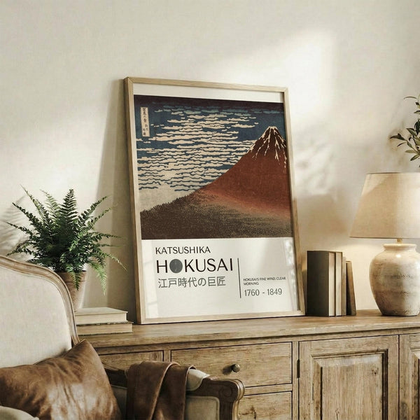 May include: Framed Japanese woodblock print featuring Mount Fuji in reddish-brown tones against blue sky with white clouds, displayed on rustic wooden sideboard alongside potted fern, ceramic vase, and stacked books in neutral-toned living room setting with beige armchair visible