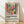 May include: Framed poster print in light wooden frame leaning against white wall on light wood floor. Artwork shows colorful still life with red flowers in orange vase, blue teapot, green pitcher, and fruit on pink tablecloth, painted in Matisse-inspired style with bold colors and simplified shapes. Below artwork is title "Celebrating Colors of Life" with descriptive text about the Matisse-inspired design.