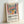 May include: Framed poster print in light wood frame leaning against white wall. Colorful still life artwork shows red flowers in yellow vase, blue and green pitchers, yellow and red fruits on pink tablecloth with vertical stripes in background. Bold, flat colors in Matisse-inspired style. Black text below reads "Celebrating Colors of Life" with descriptive paragraph about adding color to spaces.