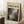 May include: Framed Jackson Pollock exhibition poster leaning against white wall beside coffee cup on wooden block and beige throw blanket. Poster features abstract black and white drip painting with "CHAOS AND CREATIVITY" and "POLLOCK 1912-1956" text on cream background in light wood frame.